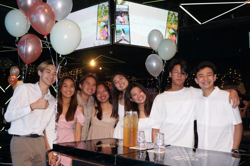 A group of young teens having fun at a Toa Payoh outlet birthday celebration, smiling and laughing together while enjoying birthday with balloons and decorations surrounding them.